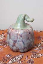 Handmade ceramic pumpkin with lavender and caramel glaze, whimsical green bent stem, on autumn patterned fabric.