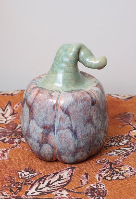 Handmade ceramic pumpkin with lavender and caramel glaze, whimsical green bent stem, on autumn patterned fabric.