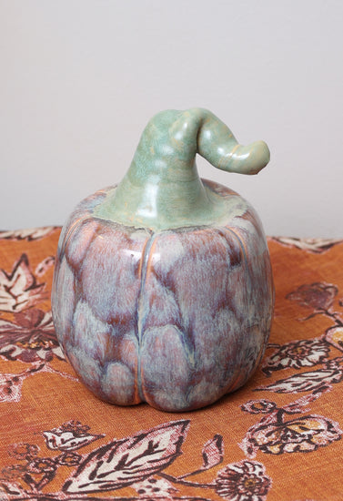 Handmade ceramic pumpkin with lavender and caramel glaze, whimsical green bent stem, on autumn patterned fabric.