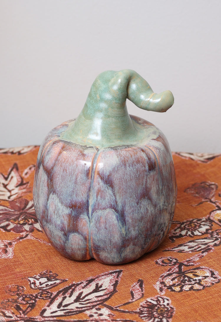 Handmade ceramic pumpkin with lavender and caramel glaze, whimsical green bent stem, on autumn patterned fabric.