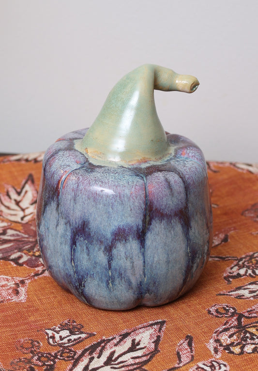 Handmade ceramic pumpkin with indigo and plum glaze, curved green stem, on fall fabric background.
