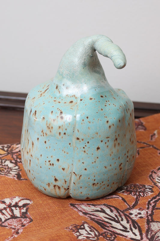Handmade ceramic pumpkin with aqua speckled glaze, curved stem, displayed on floral fall background.