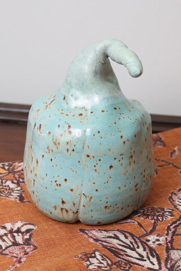 Handmade ceramic pumpkin with aqua speckled glaze, curved stem, displayed on floral fall background.