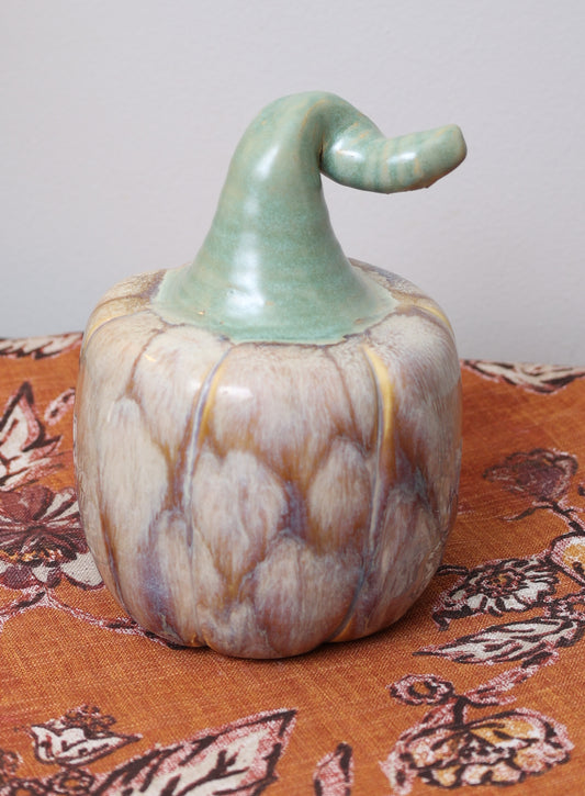 Handmade ceramic pumpkin with amber and wheat glaze, soft green curved stem, on patterned fall cloth.